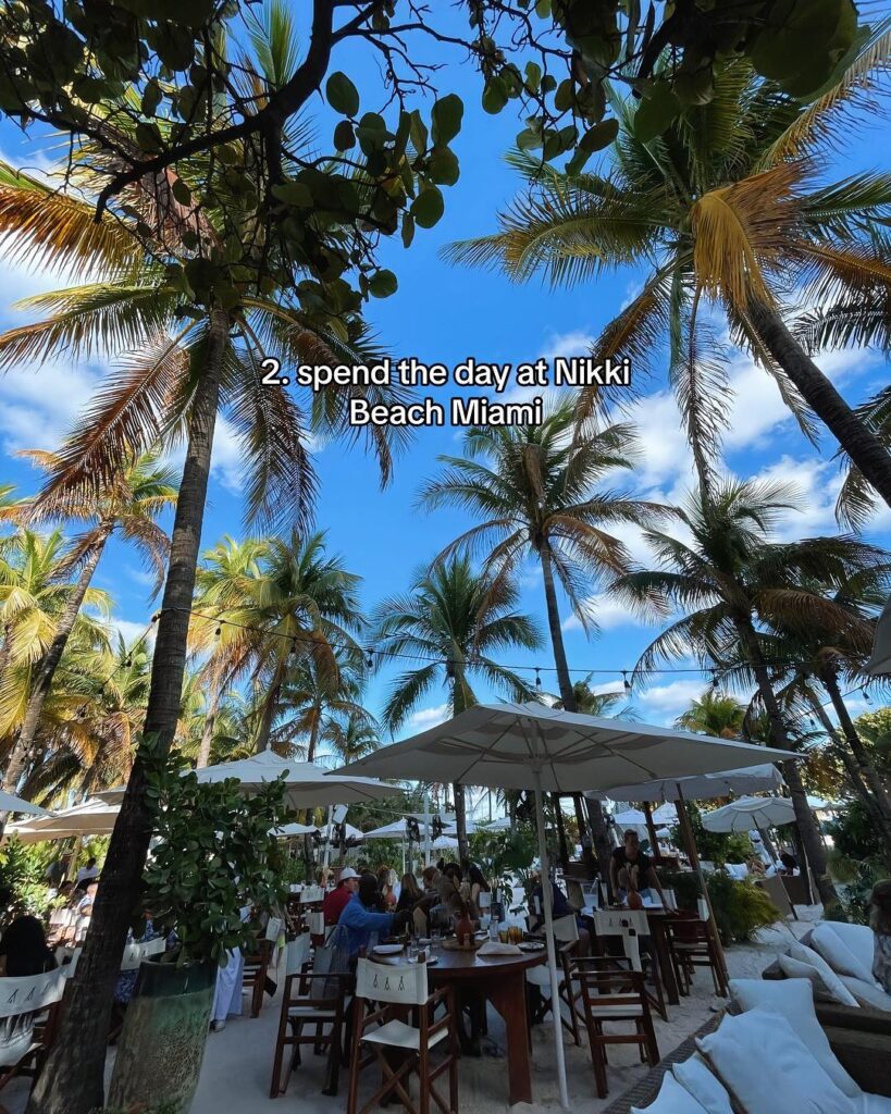 A beachside dining area at Nikki Beach with white umbrellas, wooden furniture, and swaying palm trees